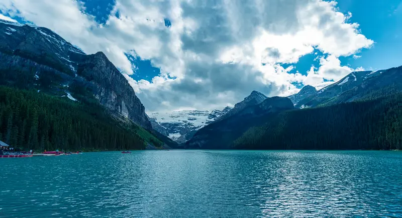 Turquoise water of Lake Louise lakeshore surrounded by pine forests and snow-capped Rocky Mountains in Banff National Park