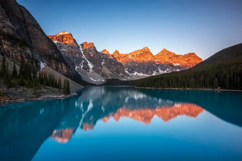 Moraine Lake at sunrise with the Valley of the Ten Peaks glowing orange and reflected in calm turquoise water in Banff National Park